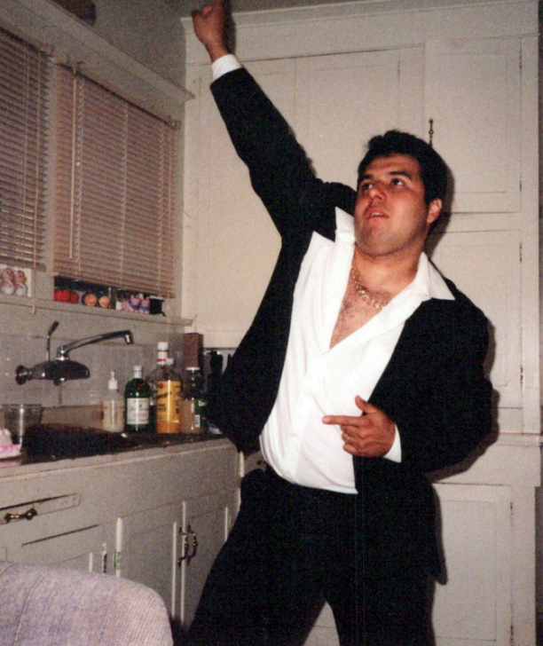 Man in disco pose wearing black suit, open shirt.
