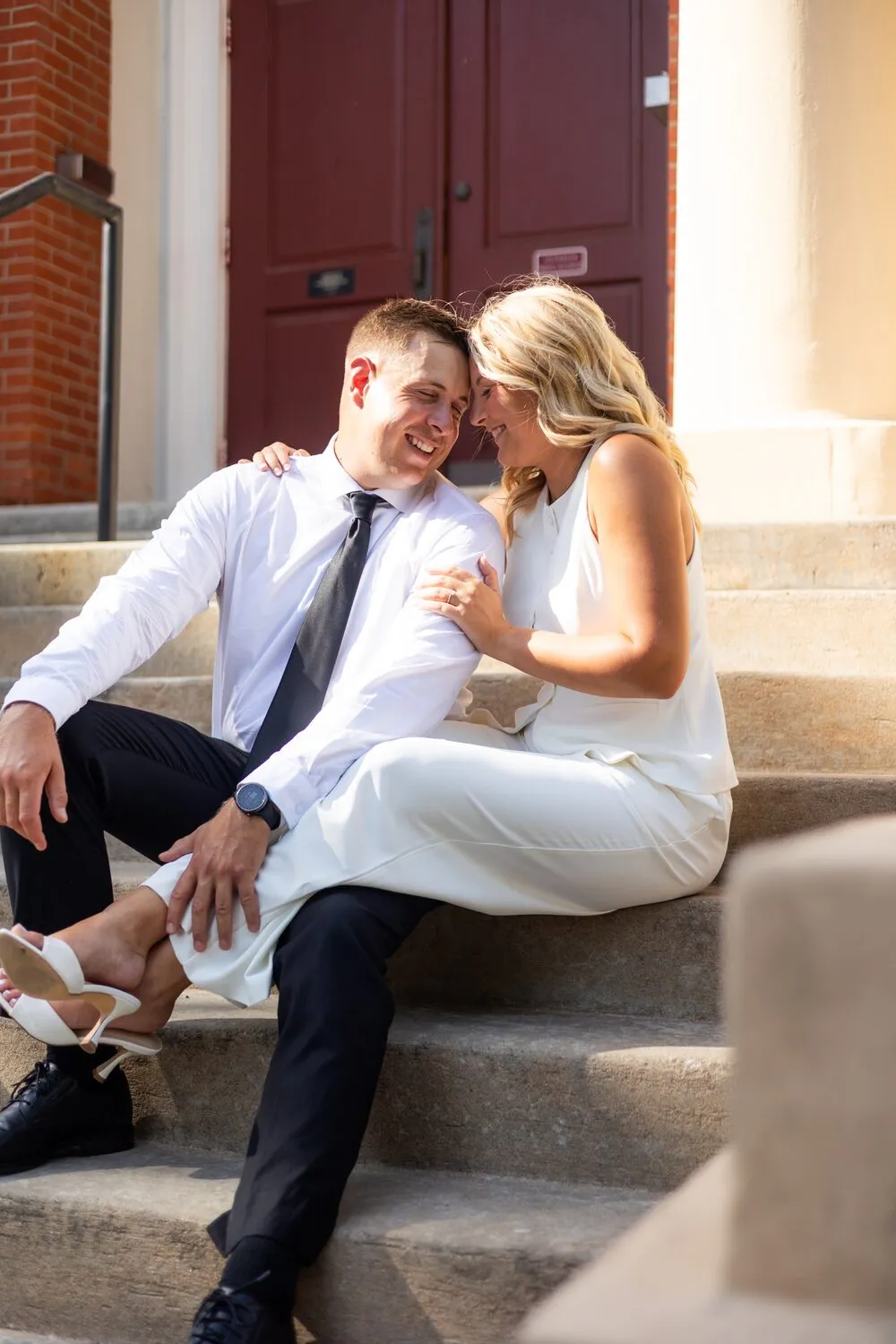 Nelius in a suit and tie and Kassidy in white, laughing on the steps by a red door